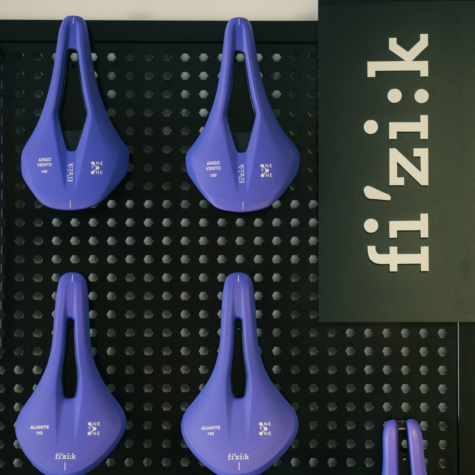Four purple bike saddles on a black pegboard with 'fi'zi:k' branding. fizik one to one custom cycling saddles.