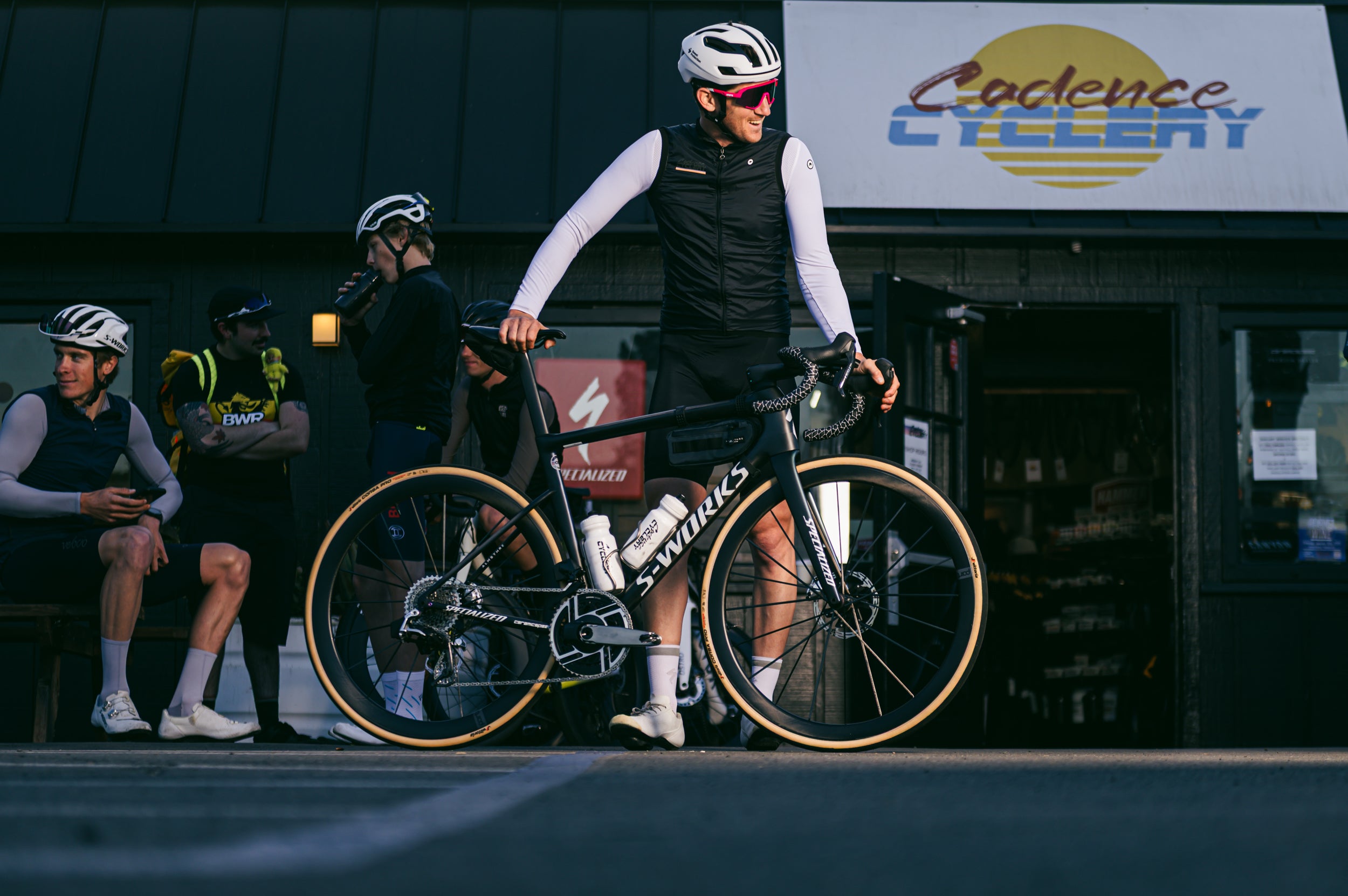 A cyclist stands behind a black Specialized road bike in front of a shop storefront, wearing cycling apparel and helmets. Other cyclists are seated nearby, and a sign for 'Cadence Cycle' is visible on the building.